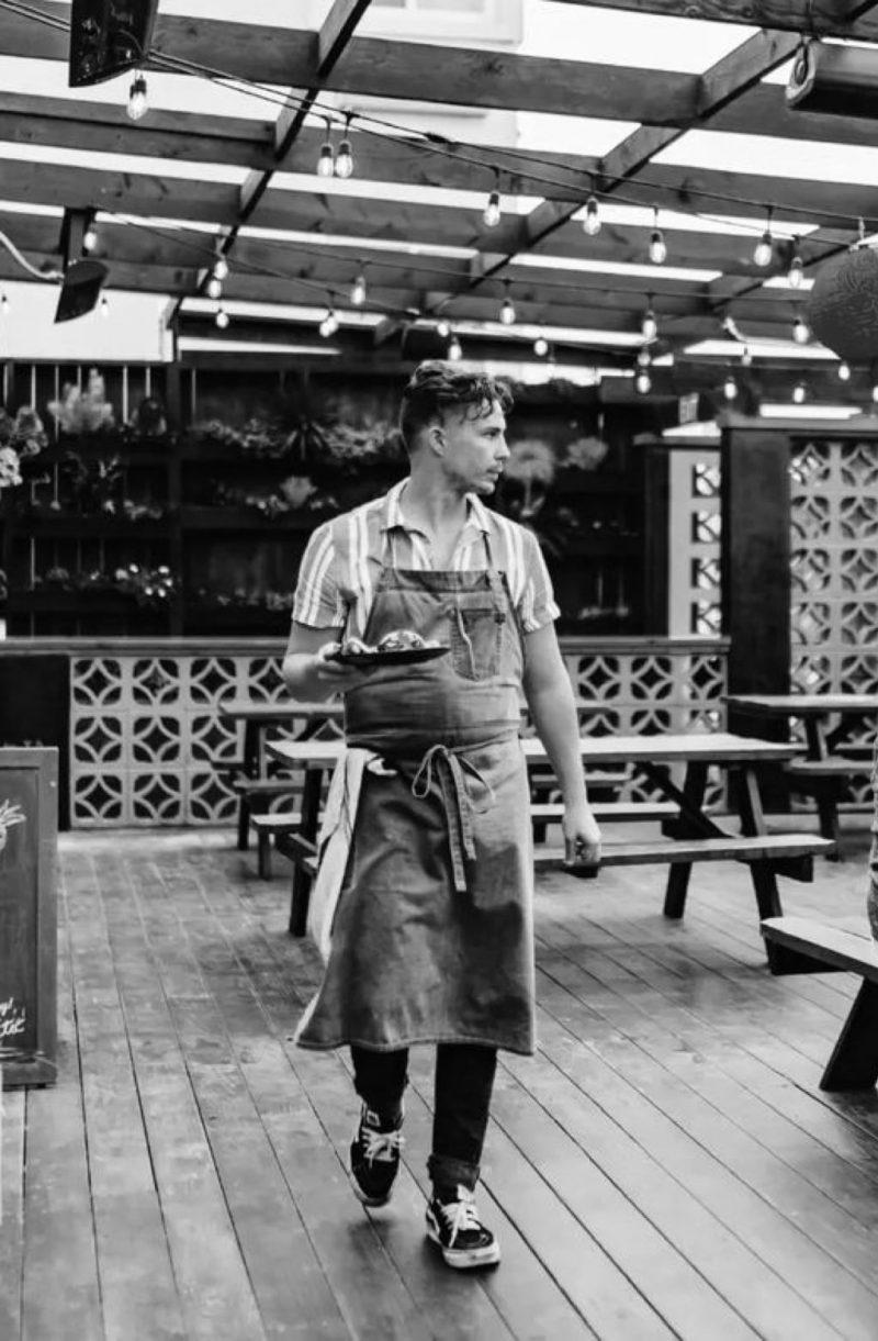 Jacob Jordan is outside in front of picnic tables and a plant wall. He is clad with an apron and fresh hair cut, walking towards the viewer with a platter of food and gazing towards his left.